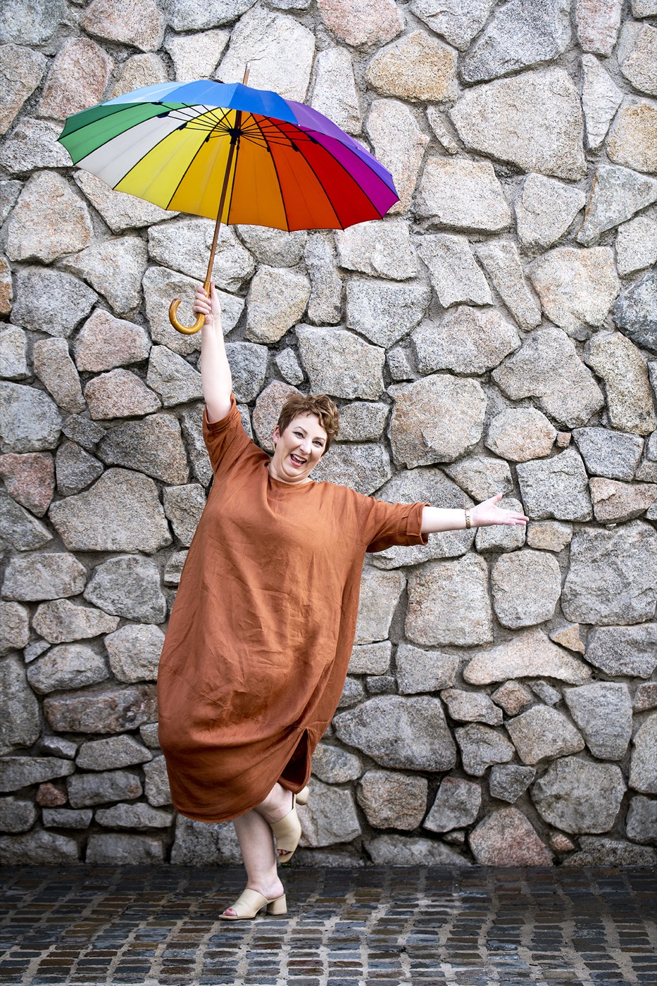Emma kicking up a heel and holding a rainbow umbrella aloft against a stone wall